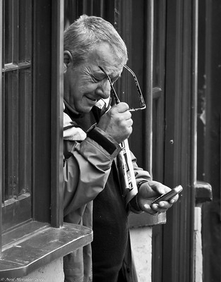 Un hombre mayor, leyendo con dificultad un teléfono móbil, mientas sostiene sus gafas en la otra mano.