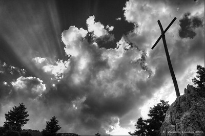 Foto en blanco y negro de una vista donde mirando hacia arriba se ven las nubes y de ellas saliendo rayos de luz, y en una esquina de la foto, la cruz de Cristo.