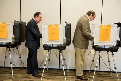 Dos cubículos de votación donde dos hombres mayores se encuentran en proceso de votación.